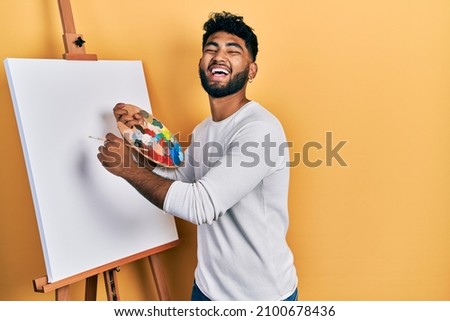 Arab man with beard standing drawing with palette by painter easel stand smiling and laughing hard out loud because funny crazy joke. 