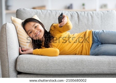 Happy asian kid pretty long-haired school-aged girl laying on couch at home, holding remote controller, watching TV alone, searching nice movie or cartoon. Kids leisure, domestic entertainment