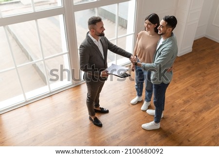 Successful Agreement, Real Estate, Home Buying Contract Concept. Happy smiling millennial buyers shaking hand with professional realtor agent in new apartment, sealing a deal together, high angle view Royalty-Free Stock Photo #2100680092