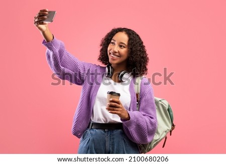 Cheerful African American female with backpack and headphones taking selfie on mobile phone, drinking takeaway coffee, photographing herself on pink studio background