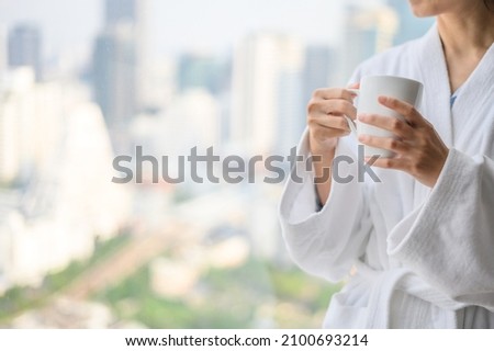 Side view of woman in bathrobe standing near the window while holding white cup of coffee and enjoy wonderful morning in the hotel, detaching from work Royalty-Free Stock Photo #2100693214