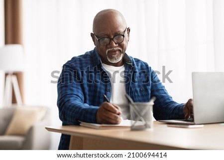 Entrepreneurship Career. Mature African American Man Taking Notes Working Sitting At Laptop Computer In Modern Office Indoors. Senior Businessman Writing Report At Workplace, Wearing Eyeglasses Royalty-Free Stock Photo #2100694414