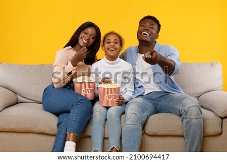 Relaxing Together. Portrait of happy young black family of three watching television, people sitting on comfortable sofa eating pop corn snack, laughing at the comedy, yellow orange studio wall