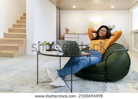 Taking Break. Smiling young black woman in glasses relaxing on bean bag chair sitting at tea table and resting, using pc laptop, happy millennial female leaning back, enjoying job, feeling pleased Royalty-Free Stock Photo #2100694498