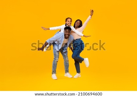 Spending Time With Family Is Fun. Full body length of excited African American man, woman and girl laughing and posing isolated on yellow studio wall. Cheerful father carrying daughter on back, banner Royalty-Free Stock Photo #2100694600