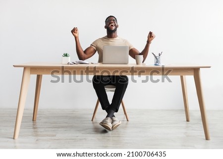 Full length portrait of excited millennial black man gesturing YES in front of laptop, celebrating success or achievement, sitting at desk in home office, copy space. Remote job, online win concept Royalty-Free Stock Photo #2100706435