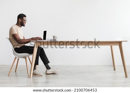 Side view of young black guy in casual wear using laptop, sitting at desk, working online from home office, copy space. Millennial African American man freelancing, having remote job Royalty-Free Stock Photo #2100706453