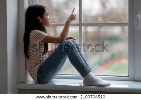 Side view of frustrated school-aged chinese girl sittng on windowsill at home, drawing on window, feeling lonely while staying home during COVID-19 pandemic, female kid missing her friends, copy space