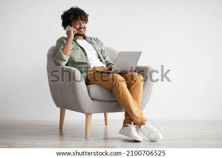 Handsome young indian guy independent contractor working online from home, sitting in comfy armchair, using laptop and having phone conversation with business partner, copy space, full length shot Royalty-Free Stock Photo #2100706525