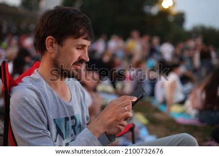 A man sits in a camping chair at an open-air music festival and looks at his phone