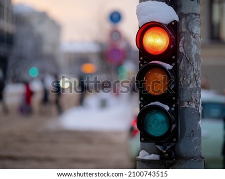 Small, bicycle traffic light section covered with snow. Red light is glowing. Blurred background.