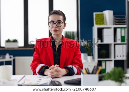 Portrait of entrepreneur with glasses in startup office. Professional businesswoman in red jacket looking confident in business workspace. Confident employee posing sitting at desk.