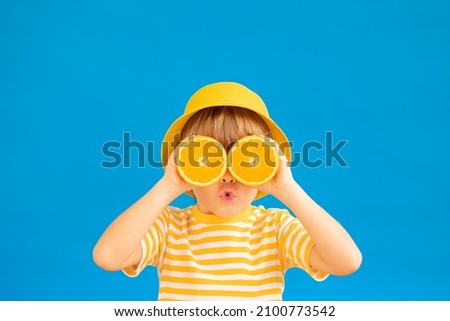 Surprized child holding slices of orange fruit like sunglasses. Kid wearing striped yellow t-shirt against blue paper background. Healthy eating and summer vacation concept