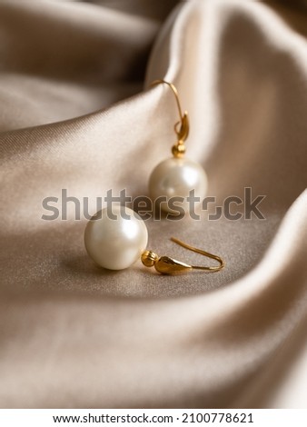 Pearl earrings with golden fittings on shiny beige silk background. Beautiful accessories for women. Elegant jewelery gift or present for wedding or saint valentine's day. Royalty-Free Stock Photo #2100778621
