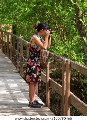 Young Short Hispanic Woman in Black Dress with Roses Takes a Picture with Compact Mirrorless Camera on a Wooden Pathway in a Mangrove