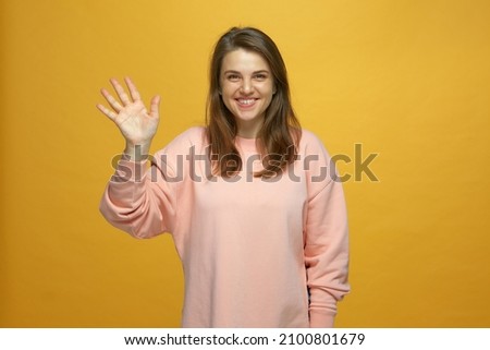Friendly young smiling girl waving hand, making hello hi welcome gesture, get acquainted on yellow studio background Royalty-Free Stock Photo #2100801679