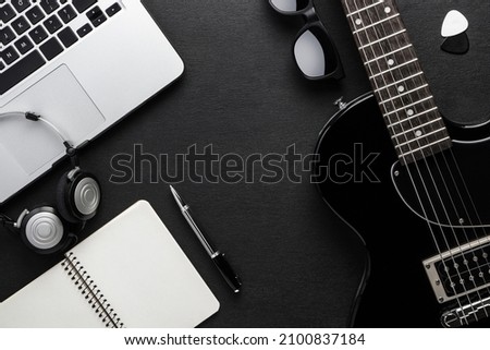 A laptop, headphones and a pen are on a black table next to a guitar, sunglasses and picks. Creative youth background concept.