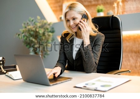 Mature happy joyful confident caucasian blonde business lady, broker or manager, sitting at table in a modern office, working with laptop, talking to client or employees on smartphone, smiling Royalty-Free Stock Photo #2100837784