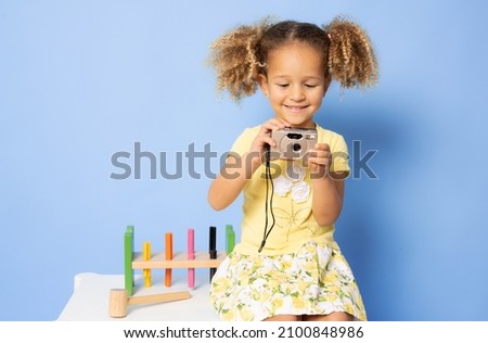 Smiling pretty little girl in dress taking photo on a camera isolated over purple background