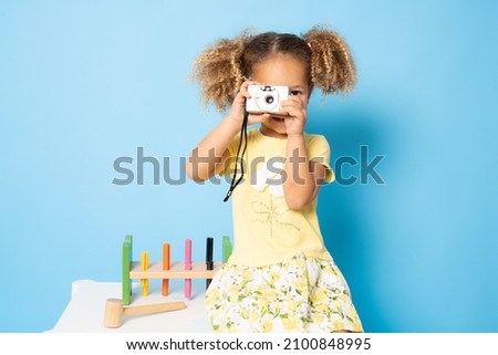 Smiling pretty little girl in dress taking photo on a camera isolated over blue background