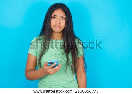 Upset dissatisfied Young hispanic girl wearing green T-shirt over blue background uses mobile software application and surrfs information in internet, holds modern mobile hand