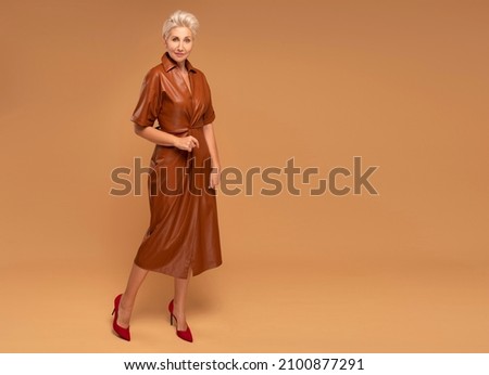 Beautiful blonde middle aged woman with short hairstyle posing in brown medium dress. Fashion photo. Elegant businesswoman. Full photo. A lot of copy space.
