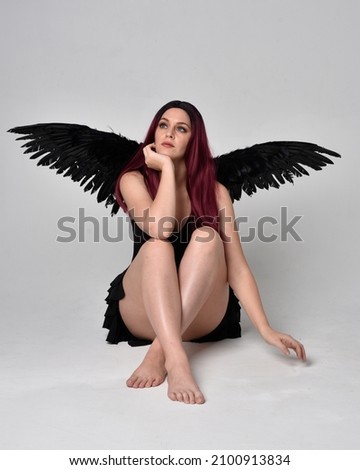 Full length portrait of pretty  female model wearing black dress and dark feather angel wings costume.  Seated pose, isolated on studio background.