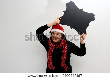 Latin adult woman with hat and Christmas garland shows banner and blank notebook to put a message of congratulations, love or wish list
