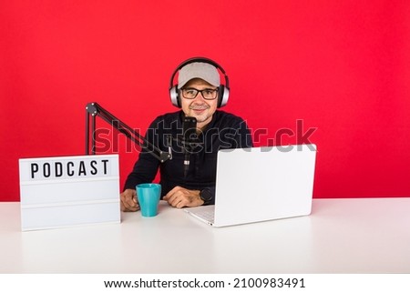 Male presenter with cap in podcast radio recording studio smiling, next to a computer, a microphone and a light box with the word podcast, on red background. Podcasting, broadcast concept