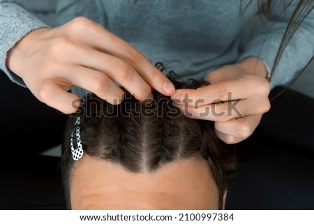 Woman is doing french braid on short men's hair, closeup view. She is doing a man's hair while sitting at home on the couch. Evening of a young married couple together. Royalty-Free Stock Photo #2100997384