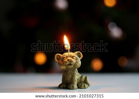 A burning candle in the shape of a tiger cub, against a background of blurry lights of a garland.