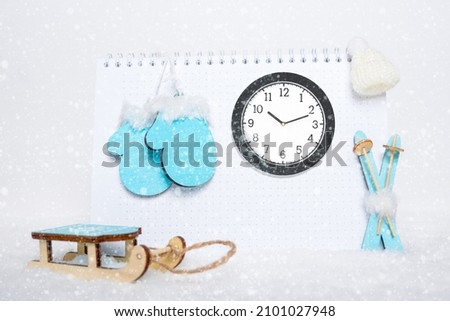 Time for winter rest. Round analog clock, toy wooden blue skis, sledges, mittens and white hat on blank notepad on snowy