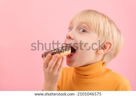 Close up portrait of child with doughnut on pink background. Towheaded boy wants to bite donut with chocolate icing.