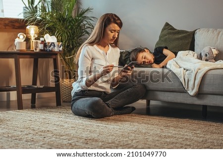 Mother checking temperature of her sick son who has thermometer. Sick child with fever and illness in bed. Boy lying sick on sofa in living room at home while his mother taking care of him