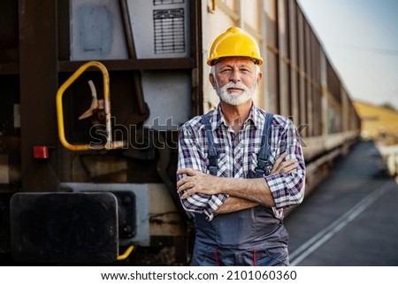 Industry transportation. A senior worker standing next to a wagon. A proud senior worker with a protective helmet on his head is standing with arms crossed next to a wagon. Royalty-Free Stock Photo #2101060360
