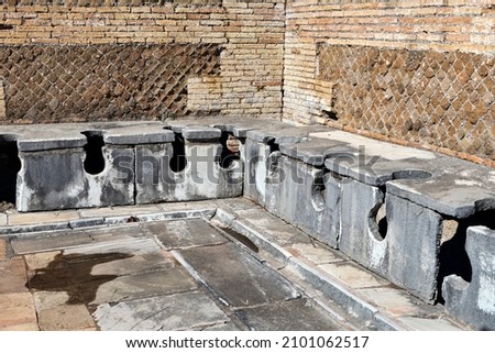 The well-preserved remains of an ancient Roman public bathroom with travertine seats and toilet drains, under the seats, the drains were constantly cleaned by a path of rough water, OstiaAntica Royalty-Free Stock Photo #2101062517