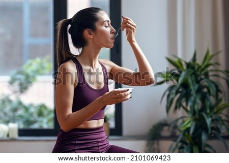 Shot of sporty young woman eating a yogurt while sitting on fitness ball at home. Royalty-Free Stock Photo #2101069423
