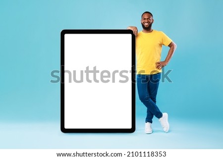 Full length portraif of cheerful young black guy leaning on giant tablet pc, demonstrating blank screen on blue studio background, free space for online advertisement, mockup for newest app