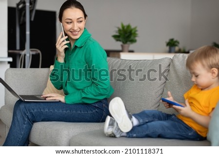 Smiling young mom working online on laptop, sitting on sofa with pc enjoying freelance job, looking little son watching cartoon on cellphone next to her
