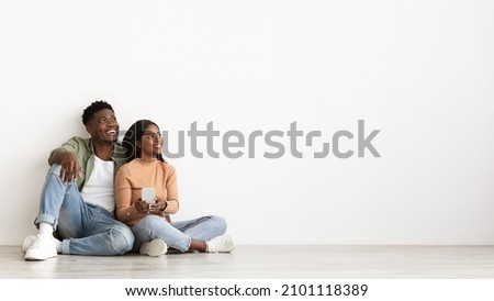 Cheerful millennial black couple with smartphone looking at copy space, sitting on floor by white wall at empty apartment, buying furniture for house online, using mobile app, panorama Royalty-Free Stock Photo #2101118389
