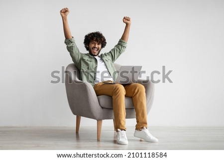 Emotional curly young hindu guy in casual sitting in armchair with modern laptop and raising hands up, celebrating success, trading on stocks and markets online, got good profit, copy space Royalty-Free Stock Photo #2101118584