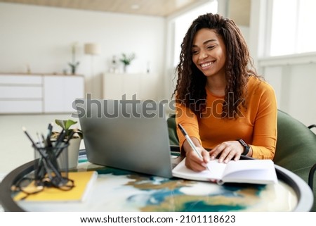 Smiling young African American woman sitting at desk working on laptop taking notes in notebook, happy millennial female studying online, watching webinar using computer and writing check list Royalty-Free Stock Photo #2101118623