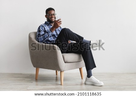 Joyful young black guy sitting in armchair with smartphone, working remotely or video chatting against white wall, free space. Millennial African American male watching movie, having online meeting Royalty-Free Stock Photo #2101118635