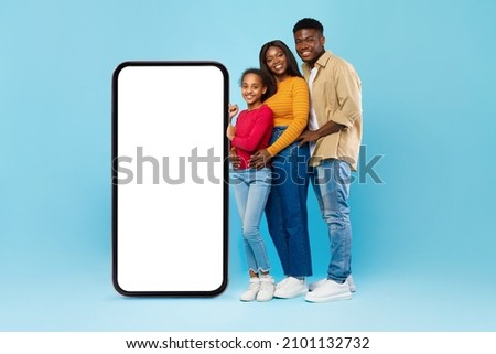 Black Family Leaning On Big Smartphone With Blank Screen Advertising Mobile Application Standing Hugging On Blue Studio Background. Great Offer, Cellular App Advertisement. People And Gadget Mock Up