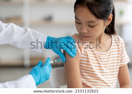 Unrecognizable doctor hands in protective gloves making vaccination against coronavirus of flu for little asian girl, home interior, closeup shot. Kids immunization against various disease concept Royalty-Free Stock Photo #2101132825