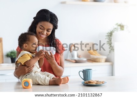 Cute Black Toddler Boy Drinking Water From Bottle While Spending Time With Smiling Young Mom In Kitchen, Thirsty Adorable Infant Baby Sitting On Counter Table And Enjoying Healthy Drink, Copy Space Royalty-Free Stock Photo #2101132867