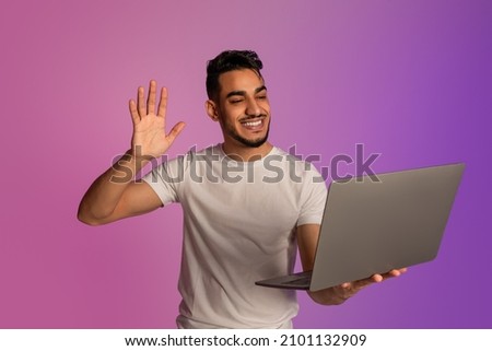 Portrait of young Arab guy waving at laptop, having video call on webcam, conferencing online in neon light. Cheerful middle Eastern man chatting on internet, enjoying remote meeting on pc