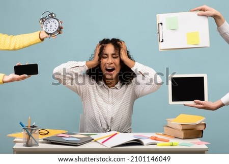 Young African American woman sitting at desk surrounded by hands with alarm clock and gadgets, screaming in despair, feeling exhausted, suffering from burnout. Multitasking, work life balance concept Royalty-Free Stock Photo #2101146049