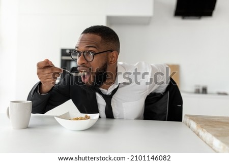 Portrait of busy African American man in glasses having breakfast in kitchen at home, wearing suit while eating cereal, rushing to office, hurry to work in the morning, being late for meeting Royalty-Free Stock Photo #2101146082