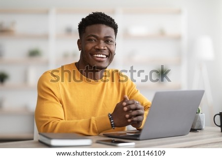 Portrait of positive young black guy in yellow sweater sitting at table, working on laptop, sending emails, office interior. Cheerful african american man freelancer working from home, copy space